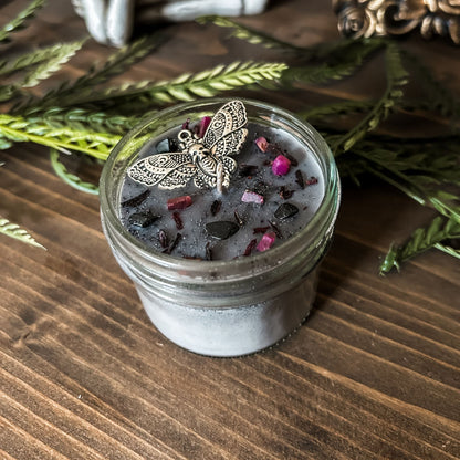 A gothic gray candle that is decorated with a gothic moth, spooky pink crystals, and dark glitter.