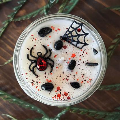 A white Halloween candle that is decorated with a black widow spider charm, a spider web charm, spooky crystals, and gothic red glitter.