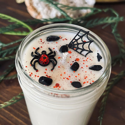 A white Halloween candle that is decorated with a black widow spider charm, a spider web charm, spooky crystals, and gothic red glitter.