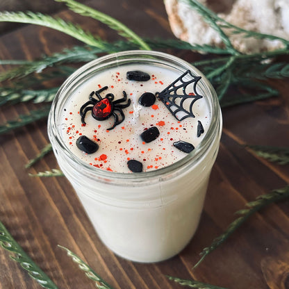 A white Halloween candle that is decorated with a black widow spider charm, a spider web charm, spooky crystals, and gothic red glitter.