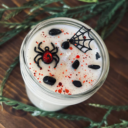 A white Halloween candle that is decorated with a black widow spider charm, a spider web charm, spooky crystals, and gothic red glitter.