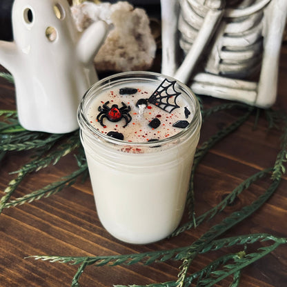 A white Halloween candle that is decorated with a black widow spider charm, a spider web charm, spooky crystals, and gothic red glitter.
