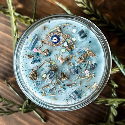 Light blue witchy candle that is decorated with a protection charm, crystals, herbs, and silver glitter to aid in the third eye chakra.