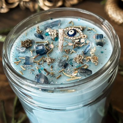 Light blue witchy candle that is decorated with a protection charm, crystals, herbs, and silver glitter to aid in the third eye chakra.