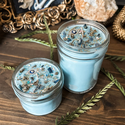 Light blue witchy candle that is decorated with a protection charm, crystals, herbs, and silver glitter to aid in the third eye chakra.