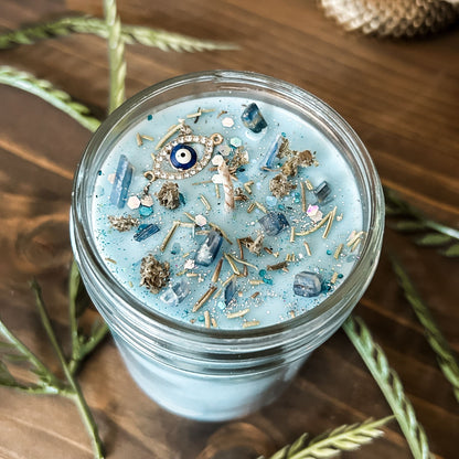 Light blue witchy candle that is decorated with a protection charm, crystals, herbs, and silver glitter to aid in the third eye chakra.