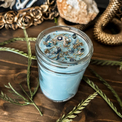 Light blue witchy candle that is decorated with a protection charm, crystals, herbs, and silver glitter to aid in the third eye chakra.