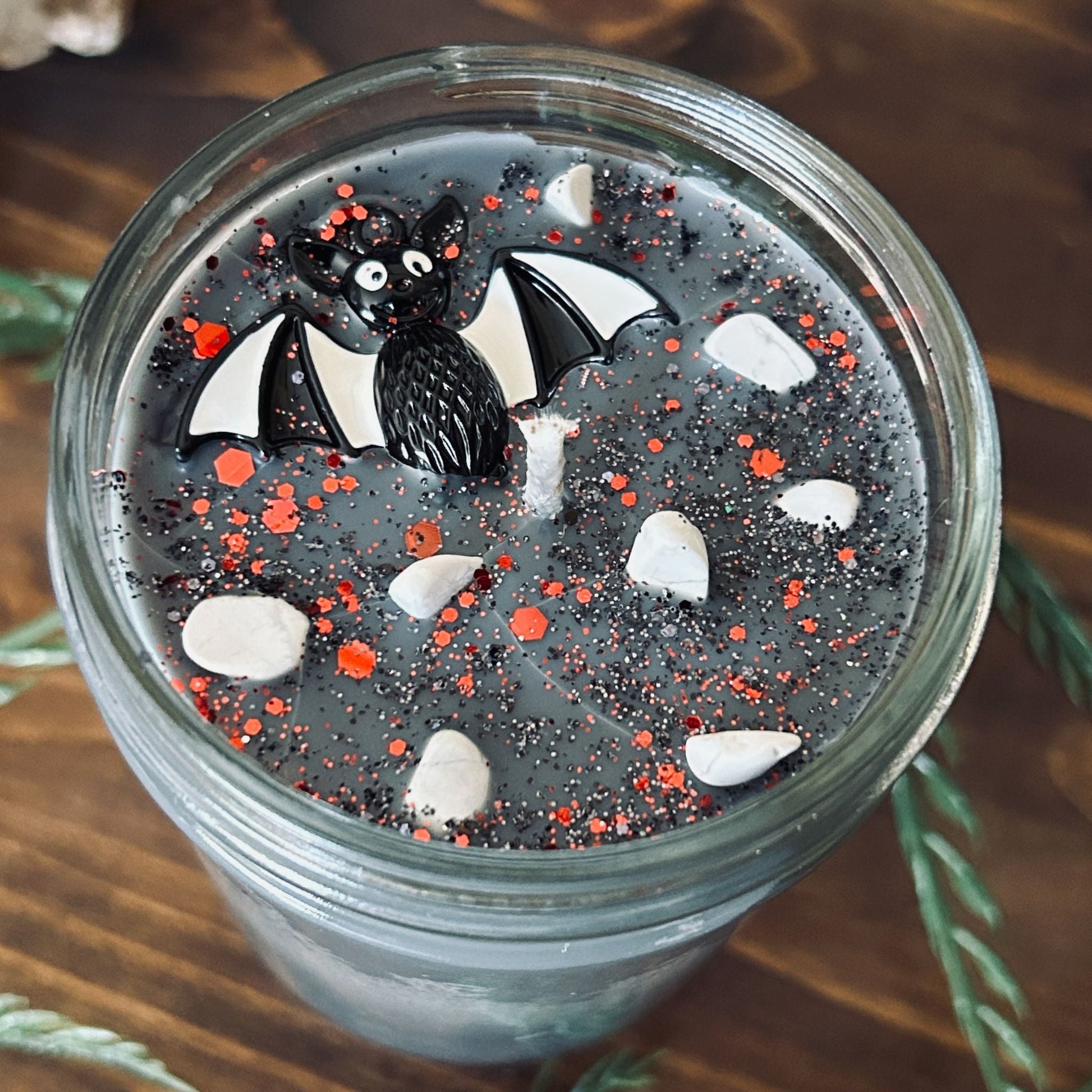 A gray Halloween candle that is decorated with a spooky black and white striped bat charm, white crystals, and gothic glitter.