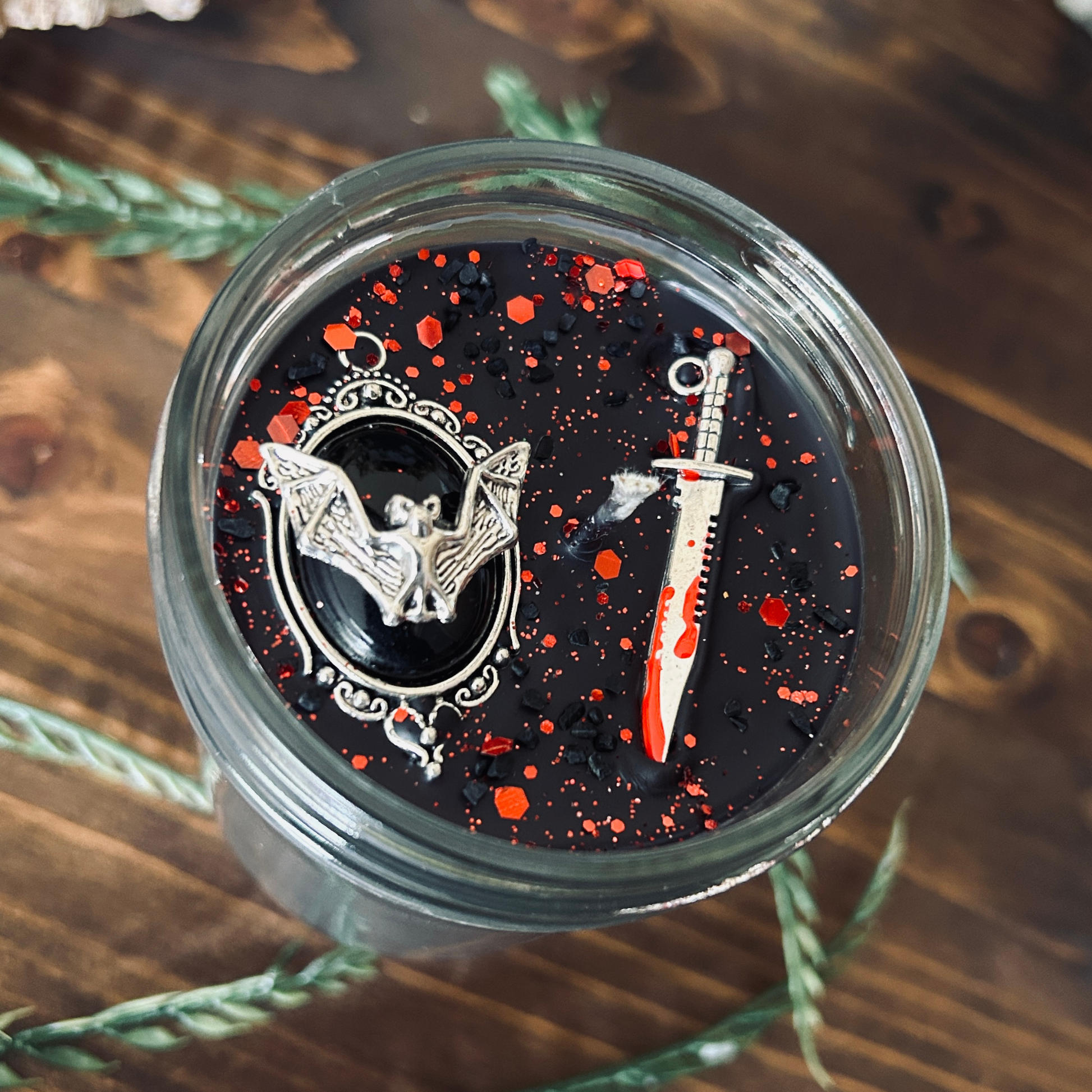 A black gothic candle that is decorated with a spooky bat, a bloody dagger, and gothic red glitter.