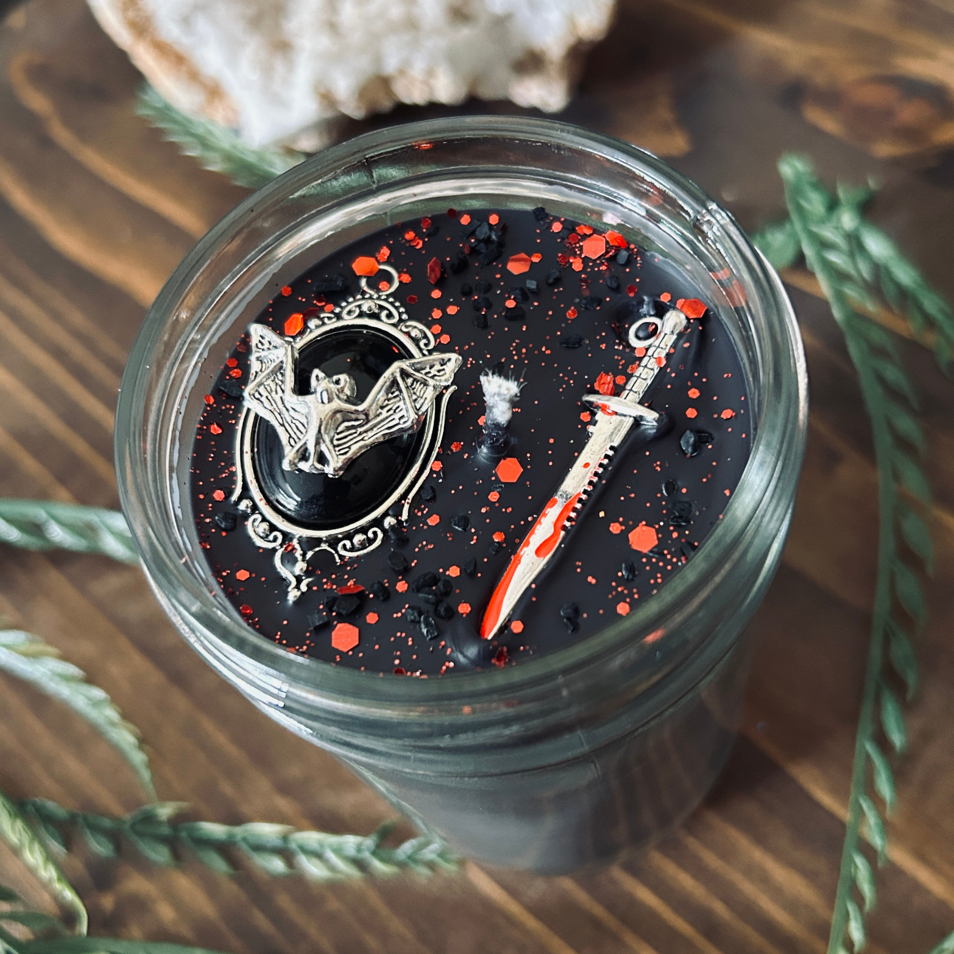 A black gothic candle that is decorated with a spooky bat, a bloody dagger, and gothic red glitter.