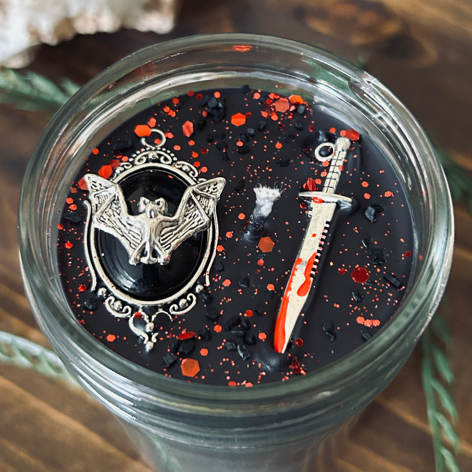 A black gothic candle that is decorated with a spooky bat, a bloody dagger, and gothic red glitter.