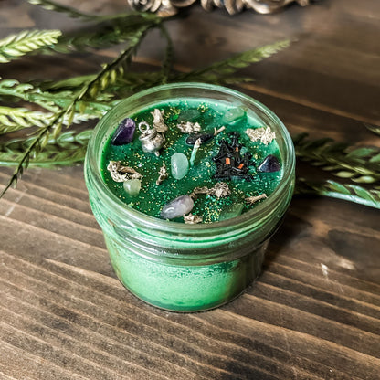 A Halloween candle that is decorated with a haunted house, a cauldron, spooky crystals, and witchy green and purple glitter.