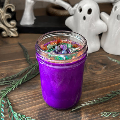 A purple Halloween candle that is decorated with a witch's broom, a witch hat, a spooky bat, witchy crystals, and magic glitter.