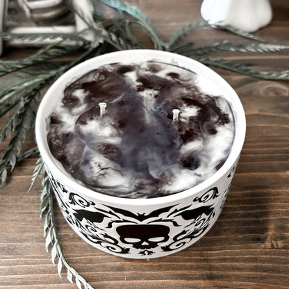 A gothic candle with a black and white swirl design that is decorated with a black skull, a raven, and a spooky bat.