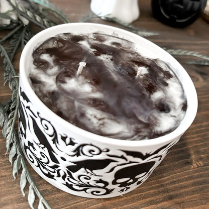 A gothic candle with a black and white swirl design that is decorated with a black skull, a raven, and a spooky bat.
